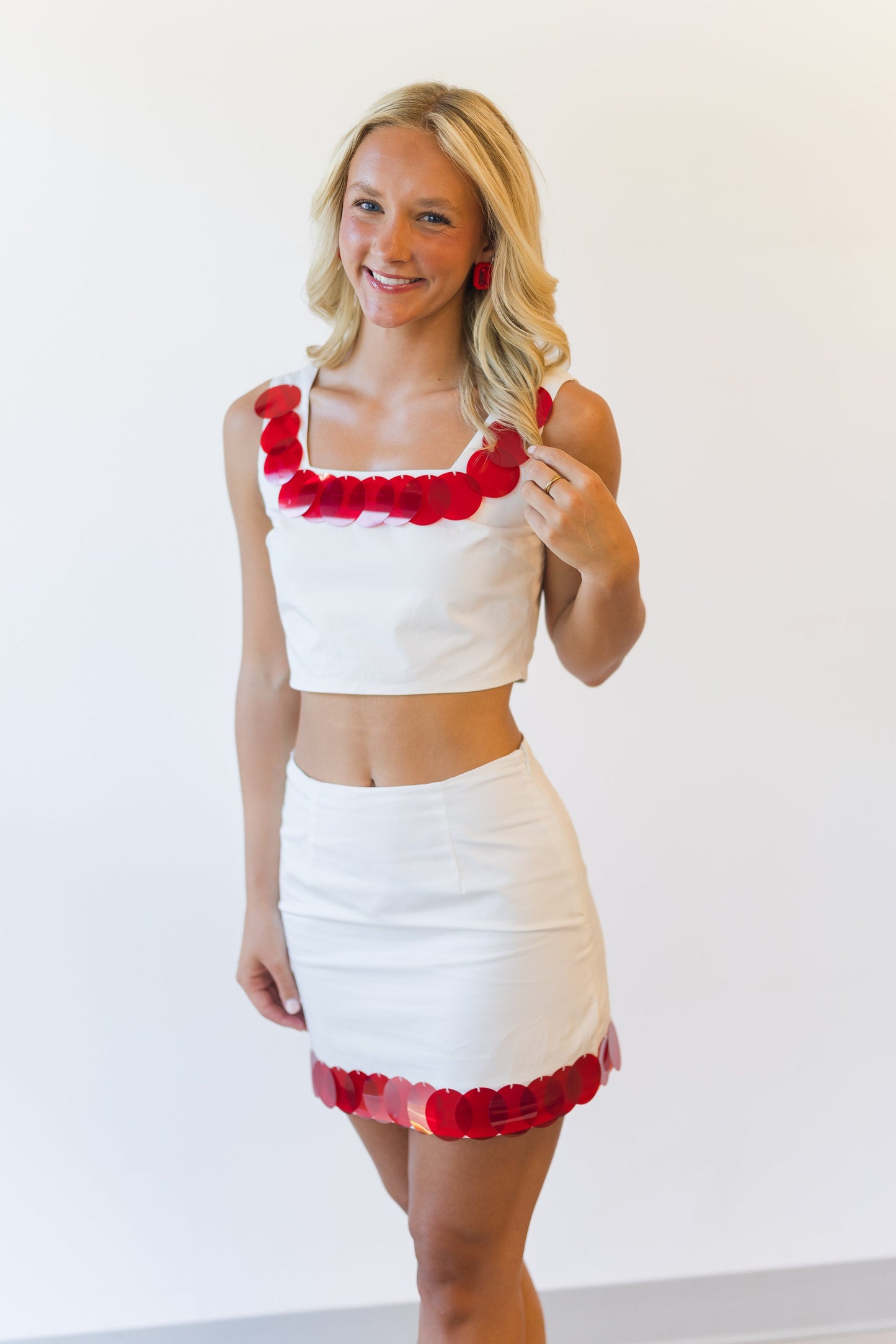 A woman modeling a white two-piece crop top and mini skirt set with red rose-patterned trim, standing against a plain white wall.