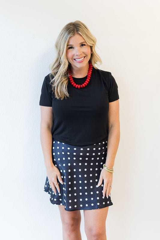 Woman wearing a black top and polka dot skirt with a red necklace against a white background